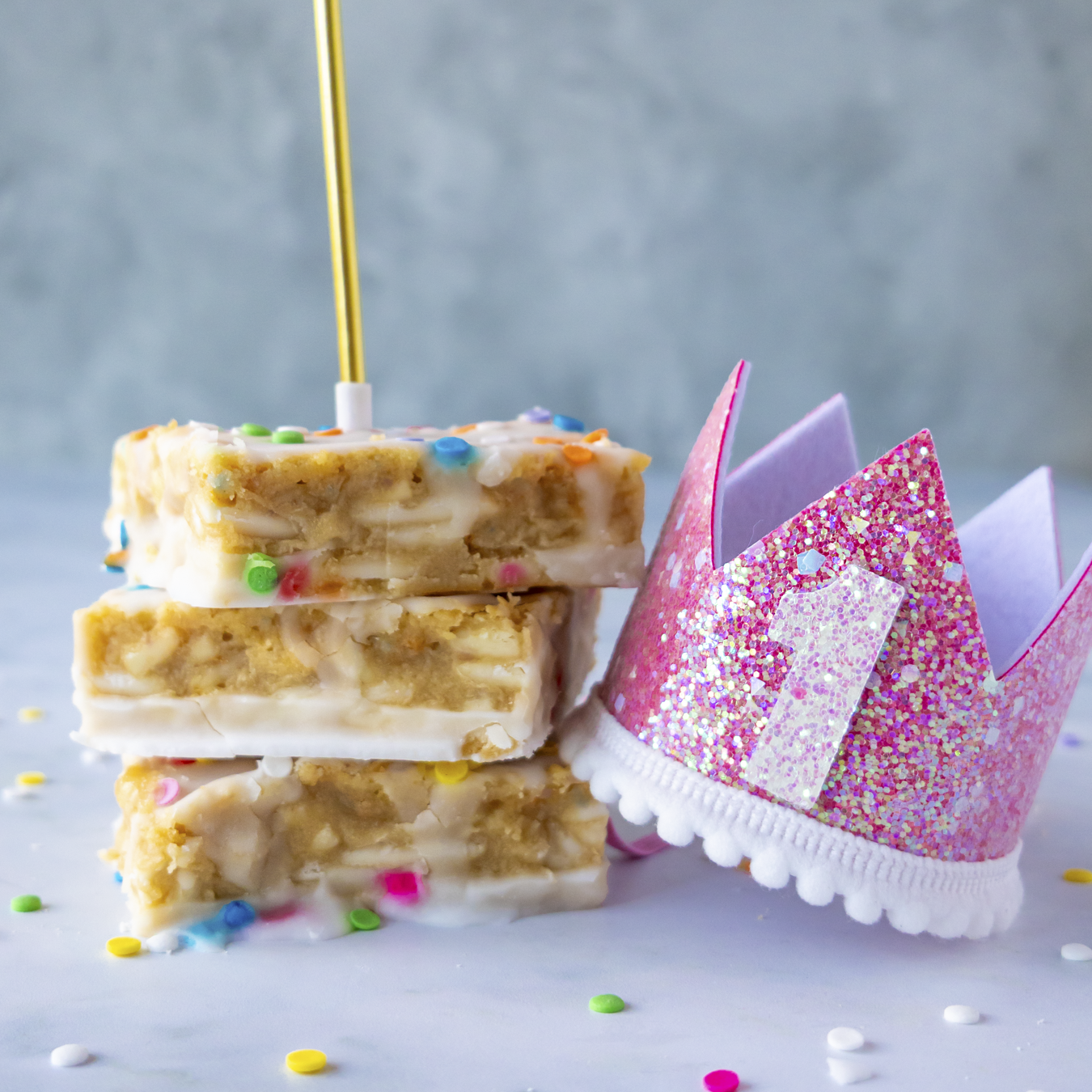 Stack of dessert blondie slab with colorful sprinkles next to a small party hat on a light gray background