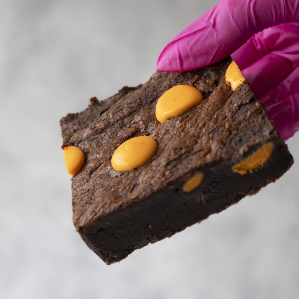 DoughGirl Brownie with orange buttons held by a gloved hand on a neutral background