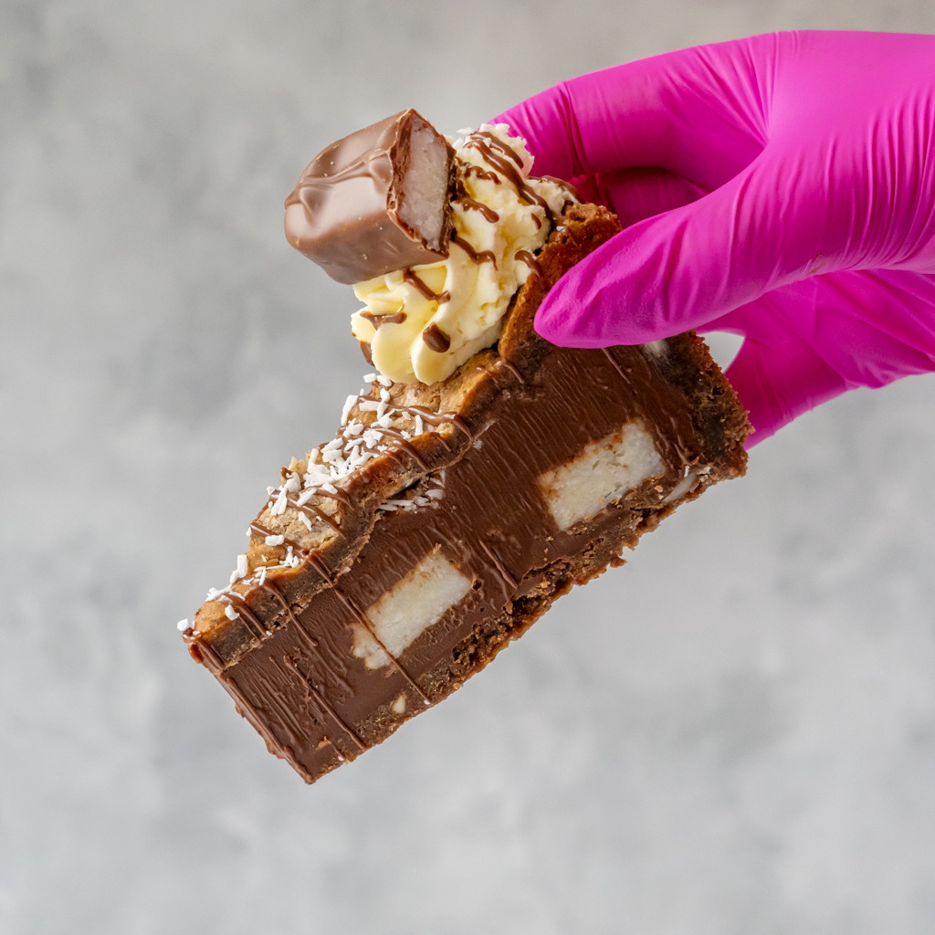 Bounty Cookie Pie with coconut flavour chocolate held by a hand wearing a purple glove on a gray background