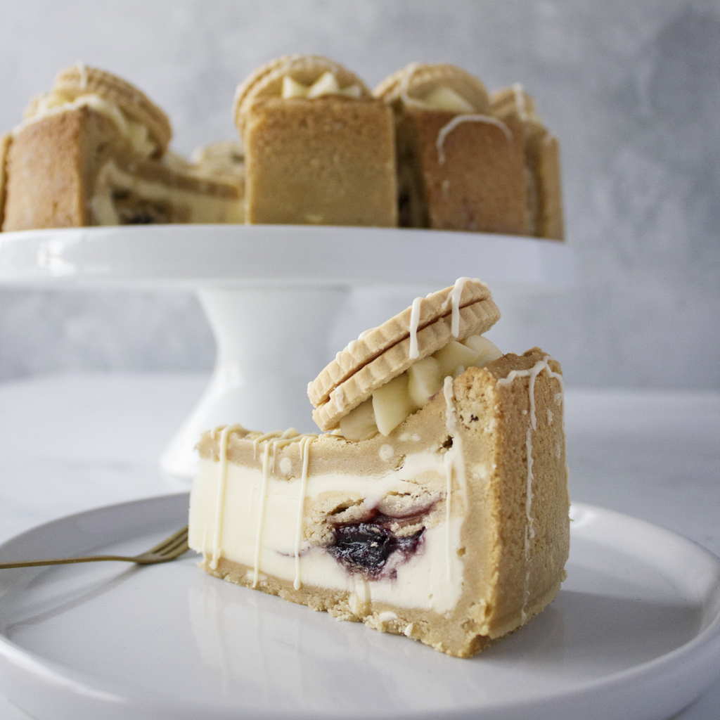 Slice of layered dessert with Jammie Dodger Cookie topping on a white plate, with a full cookie pie stand in the background.