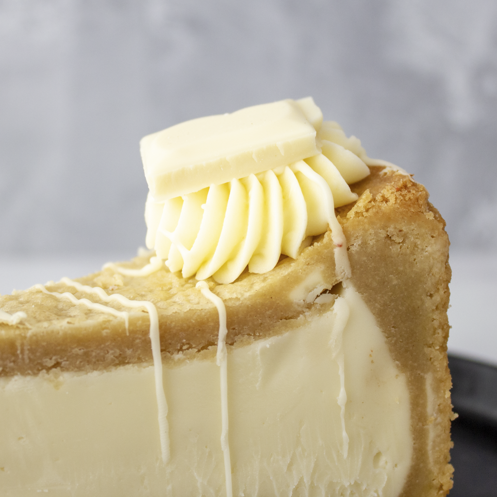 A close-up of a slice of cookie pie with Milkybar with a white choc filling, a golden-brown graham cracker crust, piped white cream on top, and white chocolate drizzle, set against a soft grey background.