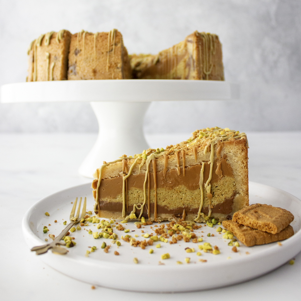 Slice of layered Pistachio Kunafa and Biscoff Cookie Pie with caramel drizzle on a white plate, with the rest of the cake on a stand in the background.