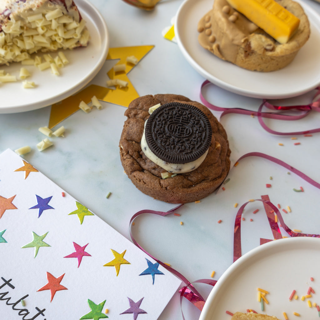 A decorated chocolate chip cookie sits on a white plate, topped with an Oreo cookie and a yellow candy bar. Other cookies and candy are partially visible around it on white plates, with confetti and pink ribbon scattered on a light marble surface.