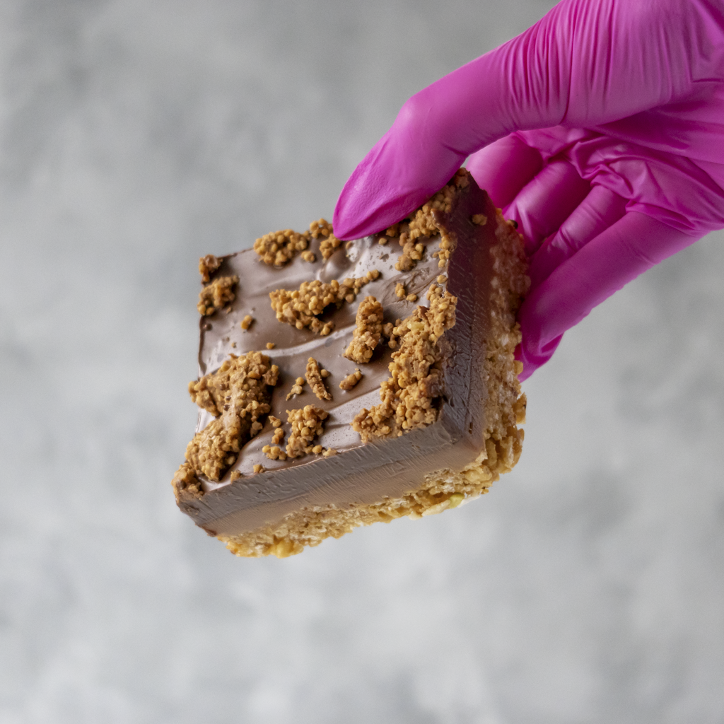 Hand wearing a pink glove holding a Cracking Caramel Crispie Slab by DoughGirl UK with Crunchy, Caramel Coated Crispies against a neutral background