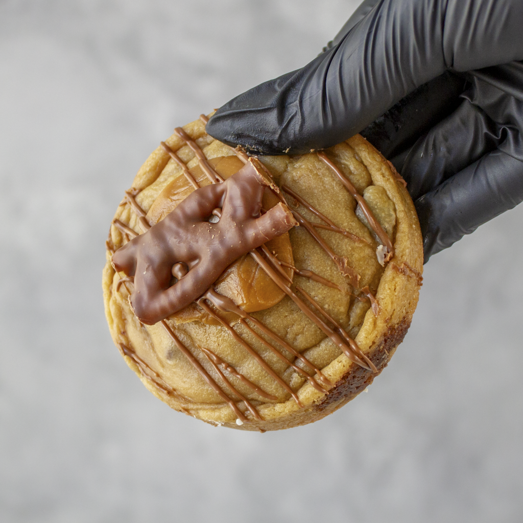 Hand holding a cookie with caramel and Curly Wurly chocolate drizzle against a neutral background
