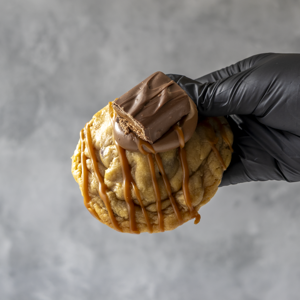Hand holding a Mars Deluxe Cookie with chocolate and caramel drizzle against a gray background