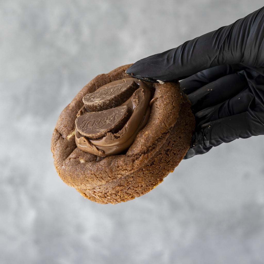 Hand wearing a black glove holding a chocolate cookie with Terry chocolate pieces on top against a gray background
