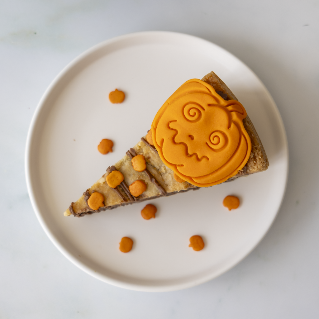 A triangular slice of pumpkin cookie pie on a white plate, decorated with bright orange pumpkin-shaped frosting and small orange sweet pieces scattered around it on a light marble surface.