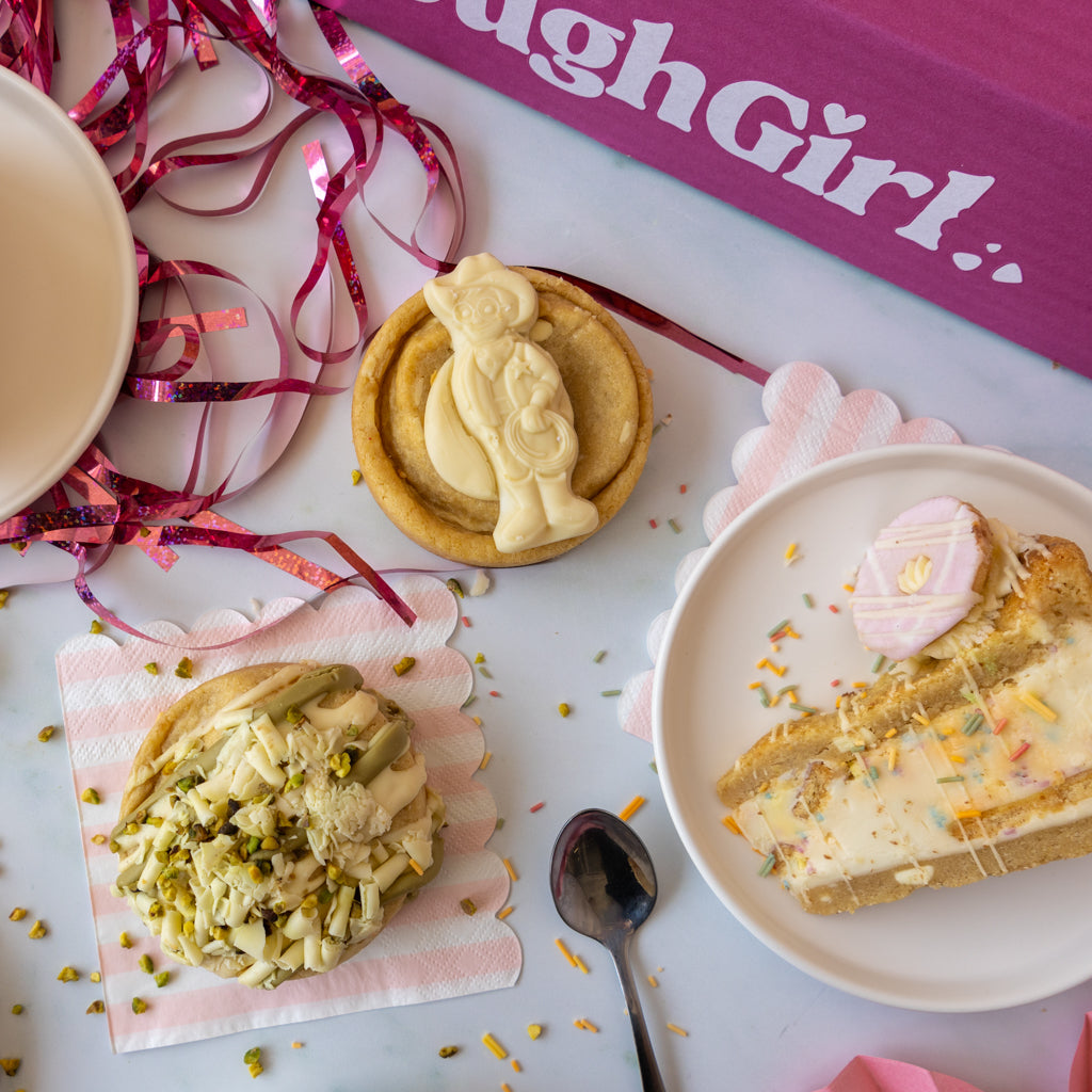 A styled flat-lay of baked goods on a white surface: a slice of layered cookie pie on a white plate, a cookie topped with pistachio pieces on pink paper, a white chocolate cookie with a cream-colored figurine decoration, pink ribbon streamers, a spoon, and a purple box with the text 'DoughGirl'.