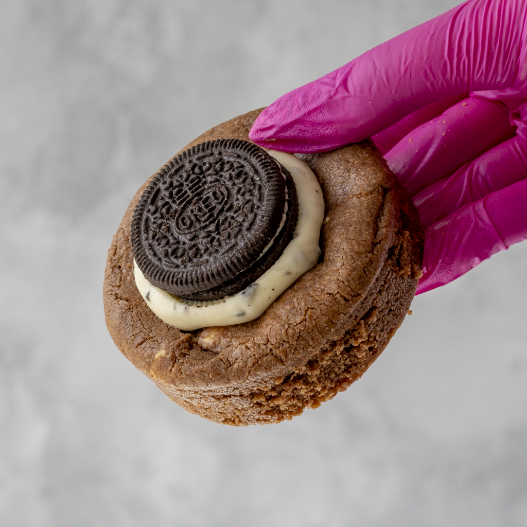Pink-gloved hand holding a thick chocolate cookie topped with cream and an Oreo biscuit
