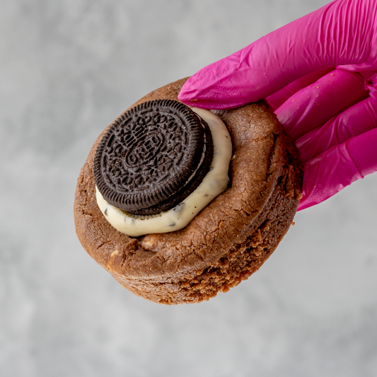 Pink-gloved hand holding a thick chocolate cookie topped with cream and an Oreo biscuit