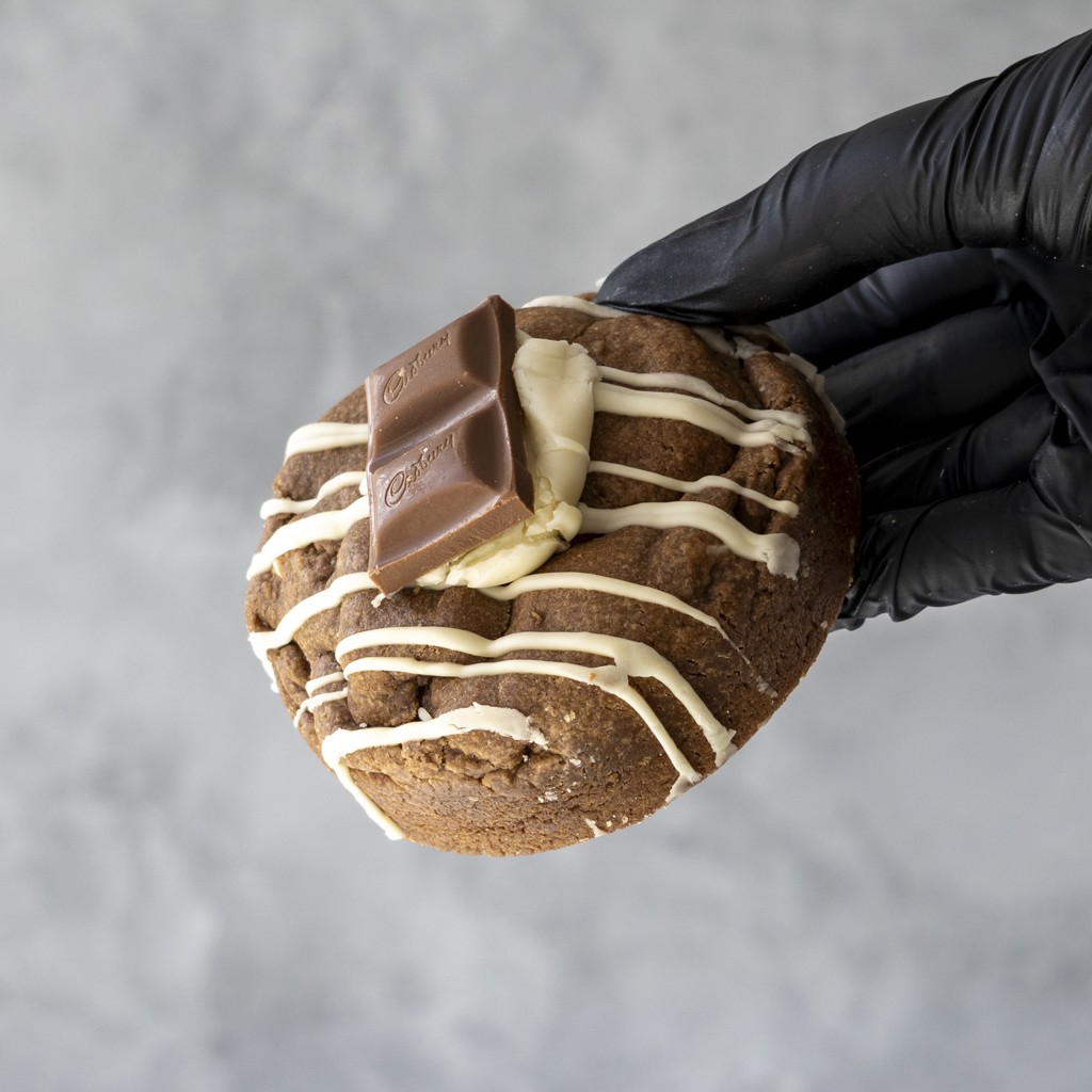 Black-gloved hand holding a thick chocolate cookie topped with white chocolate drizzle and pieces of milk and white chocolate
