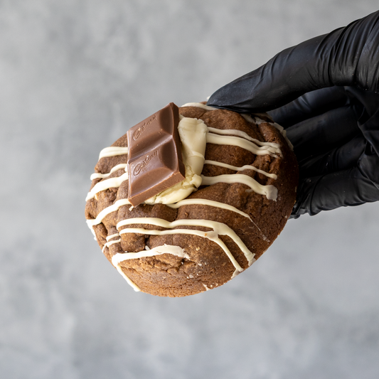 Black-gloved hand holding a thick chocolate cookie topped with white chocolate drizzle and pieces of milk and white chocolate