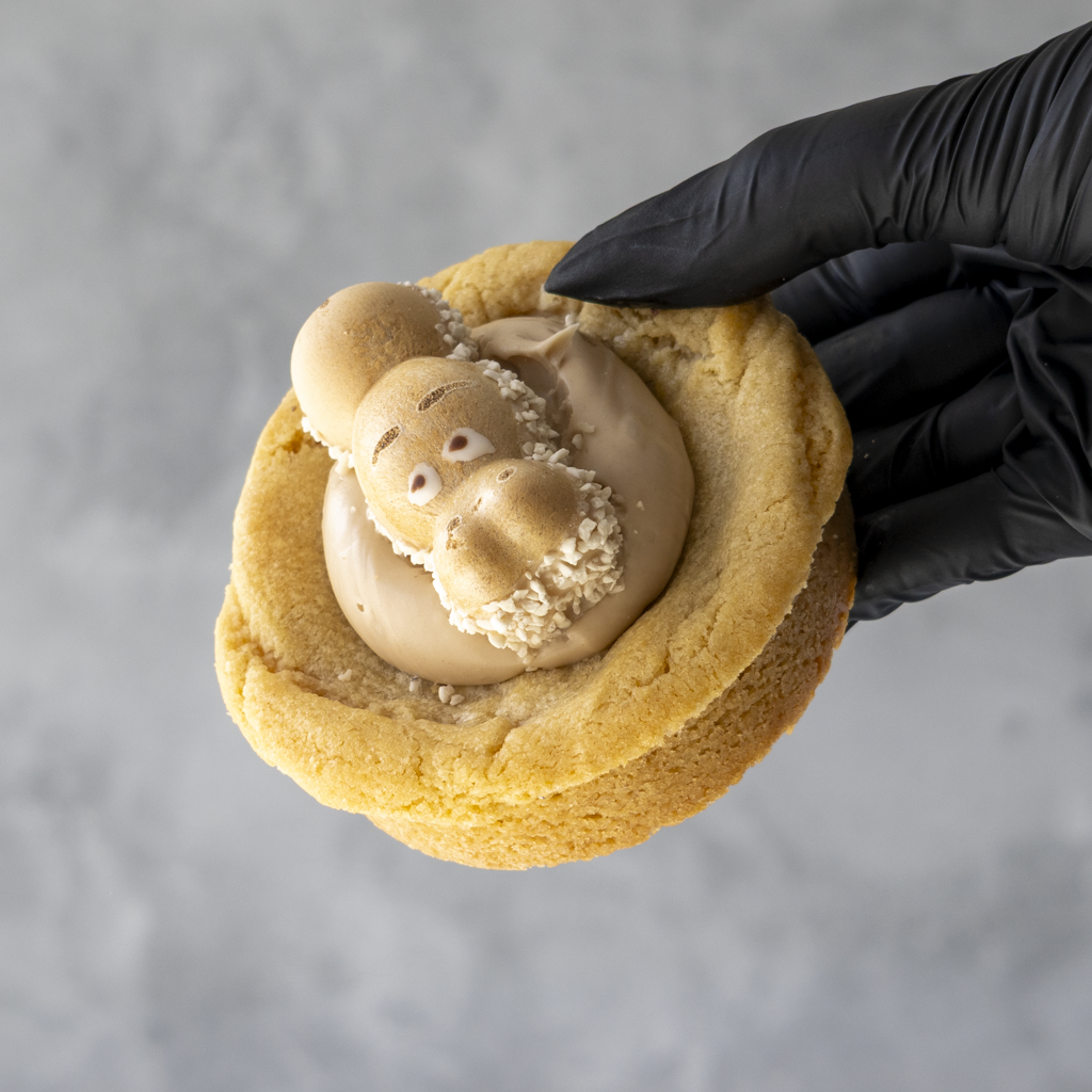 Black-gloved hand holding a thick golden cookie topped with cream and a Kinder Happy Hippo biscuit