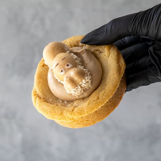 Black-gloved hand holding a thick golden cookie topped with cream and a Kinder Happy Hippo biscuit