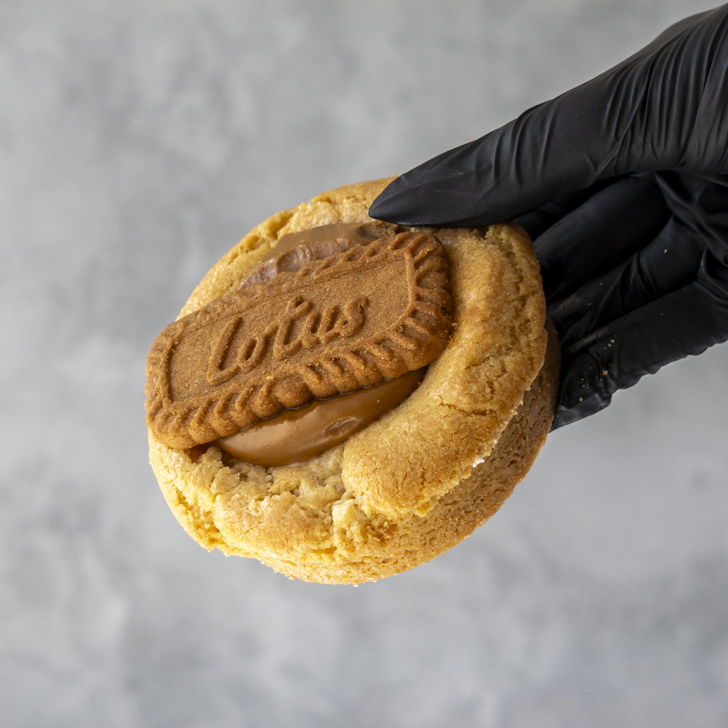 Black-gloved hand holding a thick golden cookie topped with Biscoff spread and a Lotus Biscoff biscuit