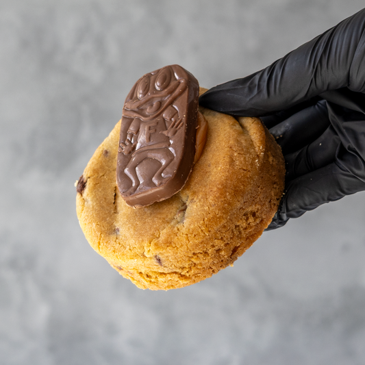 Black-gloved hand holding a thick chocolate chip cookie topped with a Cadbury Freddo chocolate frog