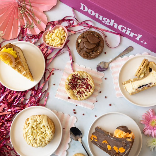 An overhead styled flat-lay of assorted baked fruity cookie an cookie pie slices on white plates, with pink tinsel, paper fans, and a pink box labeled DoughGirl.