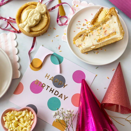 Birthday celebration spread with a birthday cake slice, decorated cookie, confetti, party hats and a Happy Birthday card.