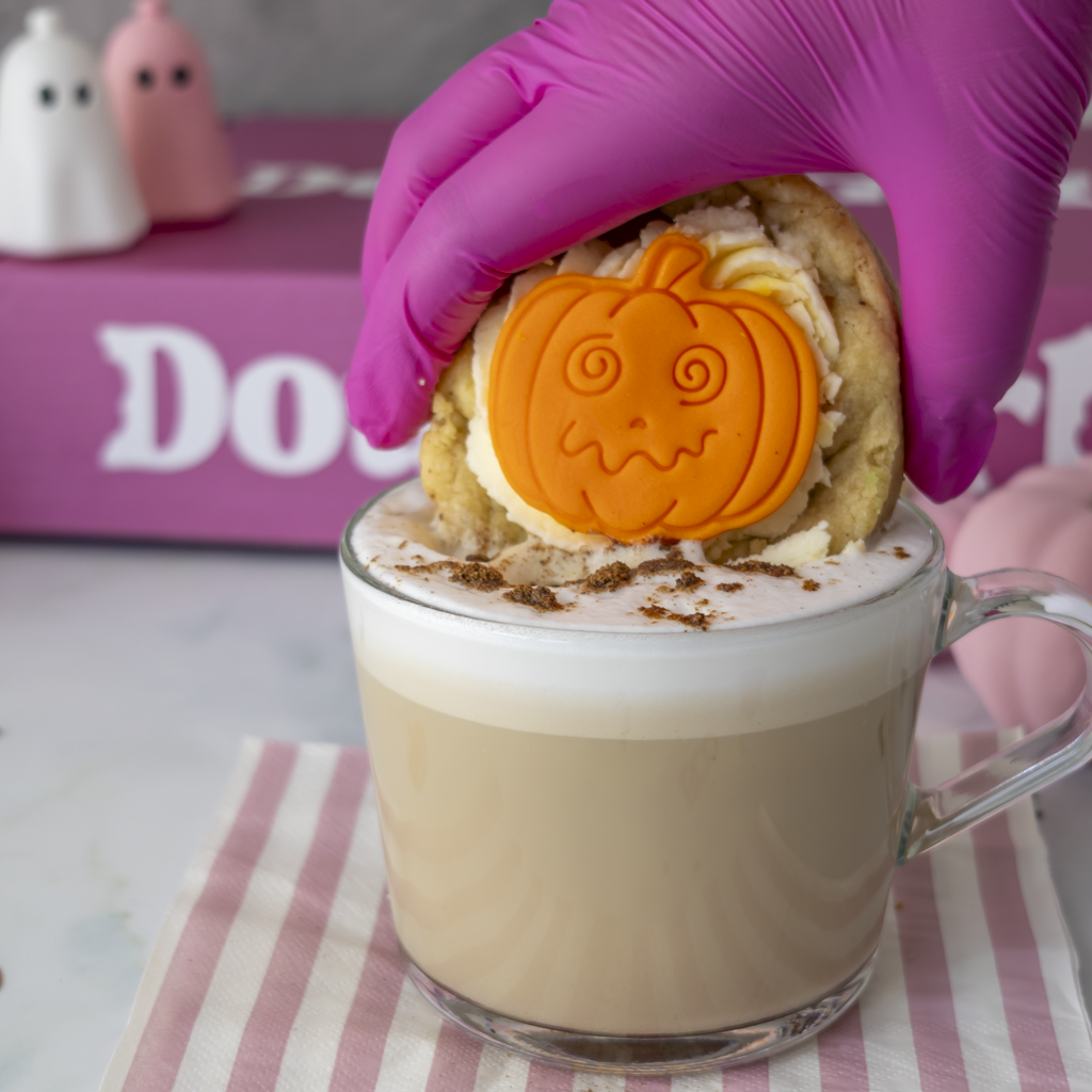 Little Pumpkin Deluxe Cookie with white frosting and an orange pumpkin decoration, served with a creamy hot drink for Halloween.