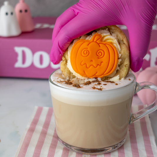 Little Pumpkin Deluxe Cookie with white frosting and an orange pumpkin decoration, served with a creamy hot drink for Halloween.