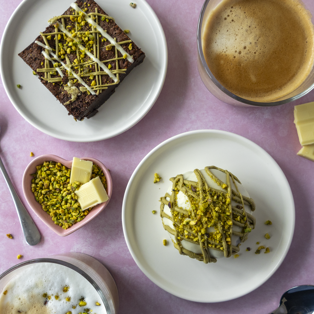 DoughGirl’s Pistachio Desserts including a choccy boom bite and a brownie slab, with coffee and other desserts, on a pink surface