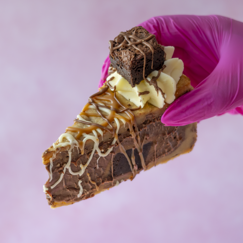 Chocolate cookie pie slice topped with brownie pieces, buttercream, caramel sauce and chocolate drizzle, held in a pink glove.
