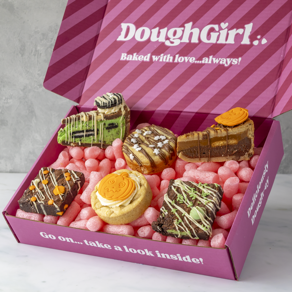 An open pink DoughGirl gift box filled with assorted baked treats, including chocolate-dipped cookies, a cookie with cream and orange frosting, photographed on a light marble surface.