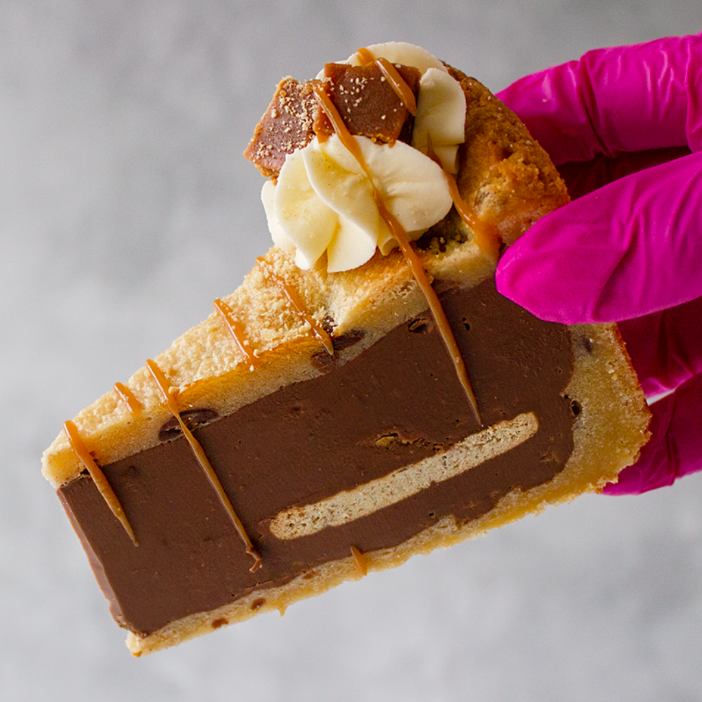 Slice of dessert with Banoffee Cookie Pie - chocolate filling, banana essence, and caramel drizzle - held by a pink-gloved hand.