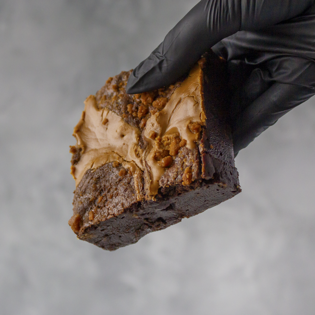 Biscoff Brownie Slab by DoughGirl with deep chocolate and spiced caramel held by a gloved hand against a neutral background