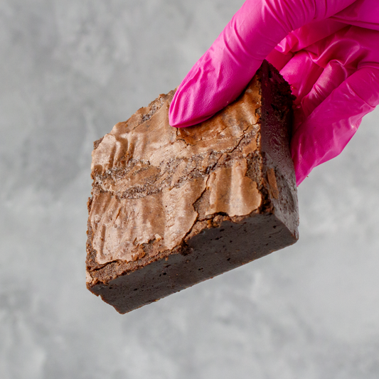 Classic Brownie Slab by DoughGirl UK held by a hand wearing a pink glove on a gray background