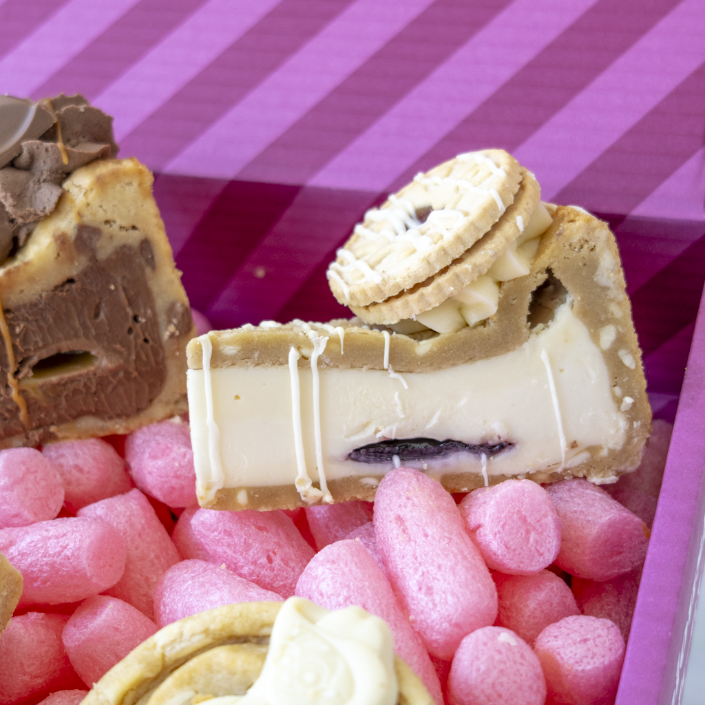 A close-up of assorted gourmet chocolate treats arranged in pink bubble wrap inside a purple box. Visible items include a chocolate-dusted pastry, a cream-filled cookie pe slice with a cookie on top, and another cookie piece with white drizzle.