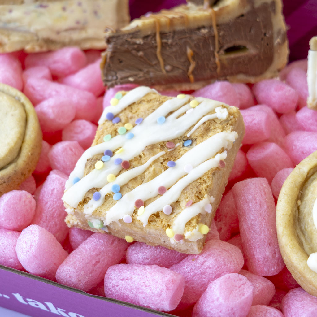 A close-up of assorted gourmet dessert bars arranged in a pink cookie box. The central bar is square, golden-brown, topped with white chocolate drizzle and multicolored sprinkles.