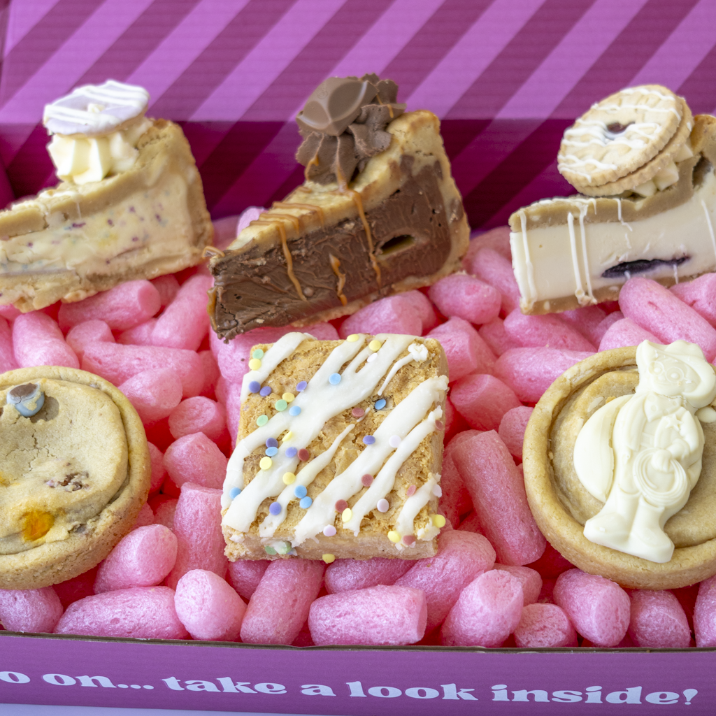 A close-up of assorted baked sweets and dessert pieces arranged in pink packing box. Visible items include a chocolate coo9kie pie slice with drizzle, a cream-topped pastry, a cookie with cream filling, a round cookie with a white figurine, and a square birthday slab with white drizzle and sprinkles.