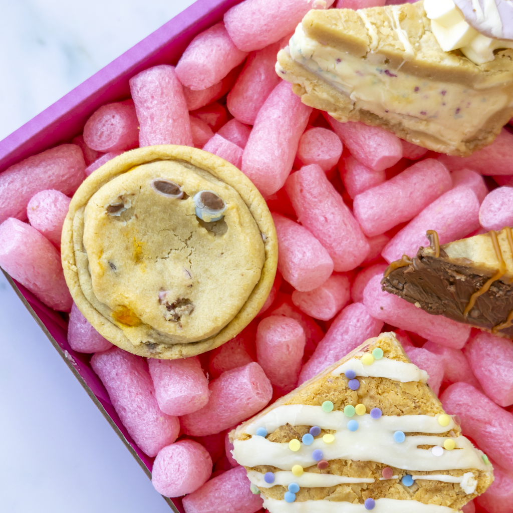 A close-up overhead view of assorted cookies arranged in a pink foam box. Visible treats include a chocolate chip cookie, a triangular cookie pie slice with white drizzle and colorful sprinkles, a layered cookie, and a chocolate-dipped cookie piece.