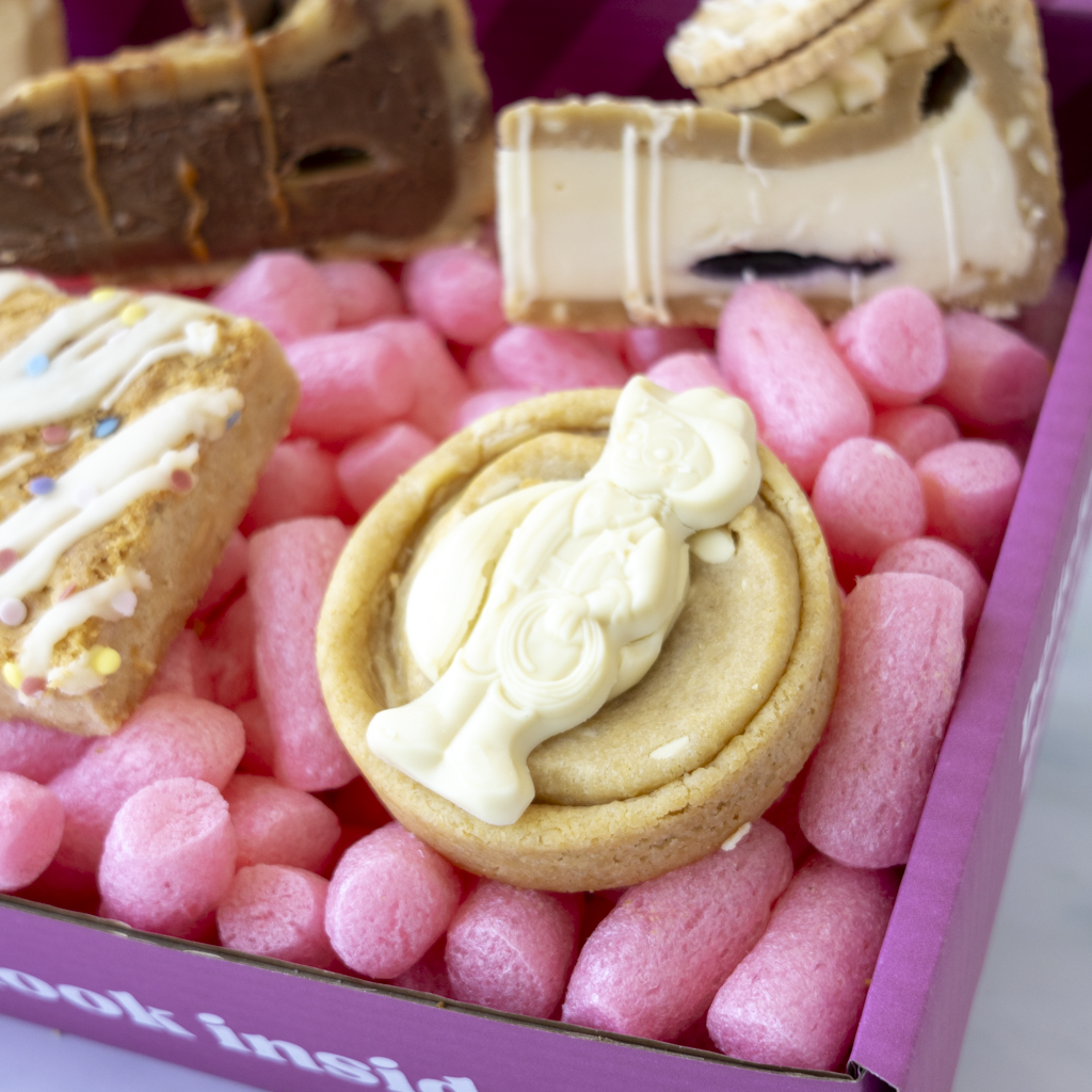 A close-up of a boxed treat assortment. In the centre is a round golden cookie topped with white cream, surrounded by other assorted sweets and cookies inside a pink box.