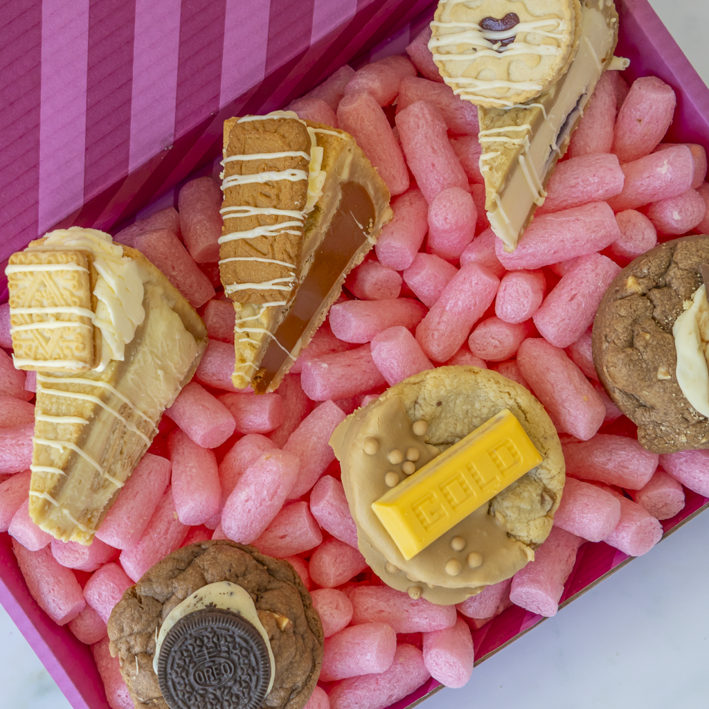 A close-up overhead view of assorted sweet treats arranged in a pink box. The treats include several cake slices with drizzles, cookies, and a chocolate bar, photographed from above inside the box.