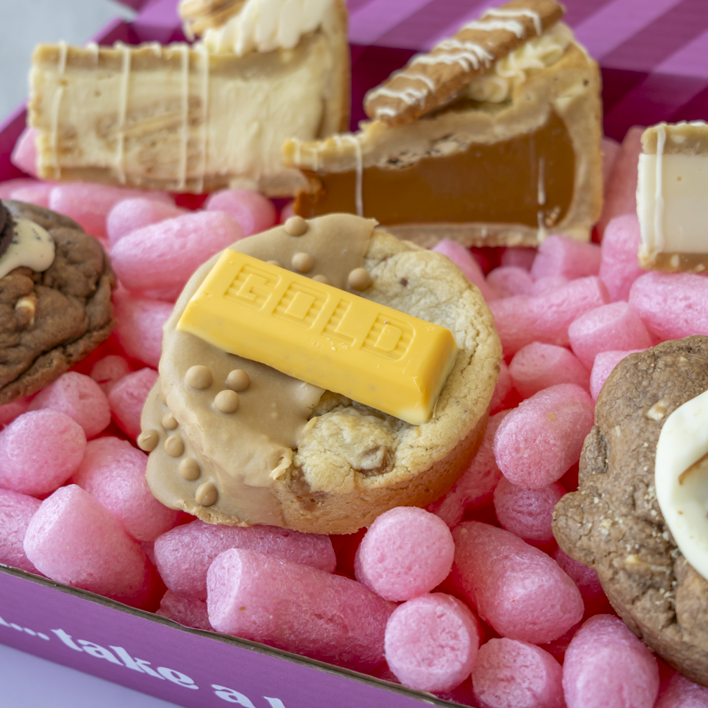 A close-up of assorted gourmet cookies arranged in a pink packing box. The center cookie is topped with a yellow chocolate gold bar and beige frosting, surrounded by pink bubble wrap and other decorated cookies including pie slices and chocolate drizzles.