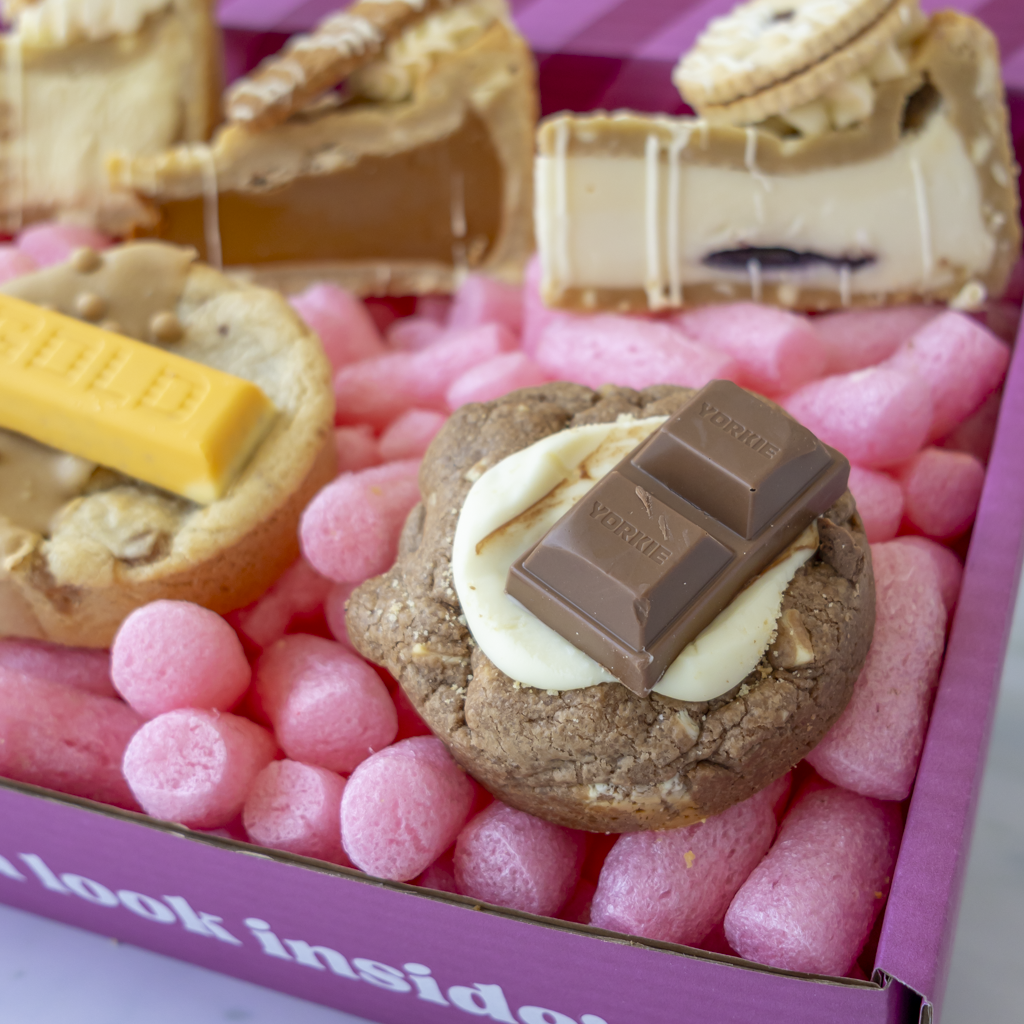 A close-up of assorted baked treats arranged in a pink cookie box. The centre item is a cookie topped with a white chocolate swirl and a dark chocolate bar embossed with 'YORKIE'.