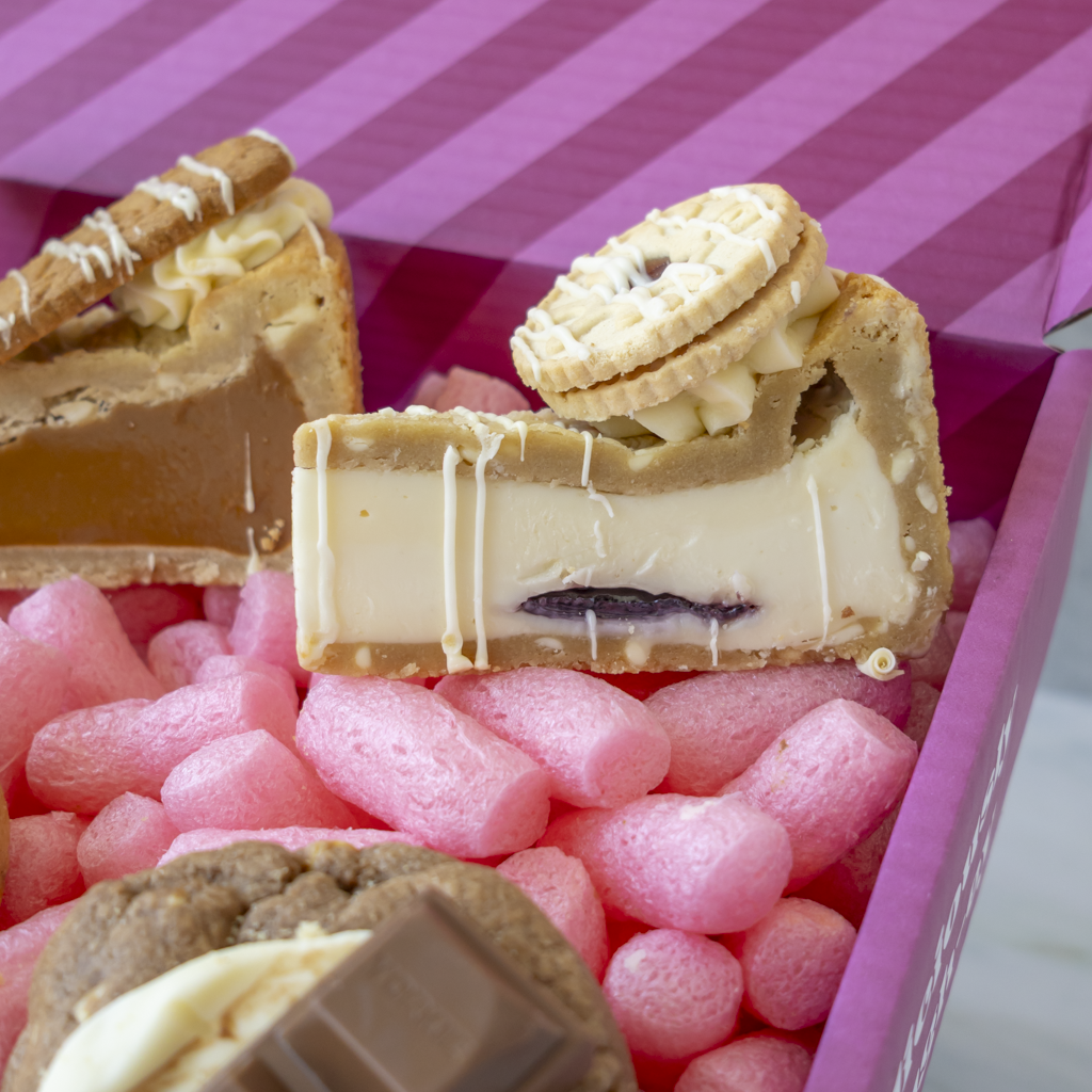 A close-up of assorted gourmet chocolates and cookies presented in a pink box. The centre chocolate piece is cut to show a creamy white filling, a dark jam centre, and a Jammie Dodger biscuit on top.