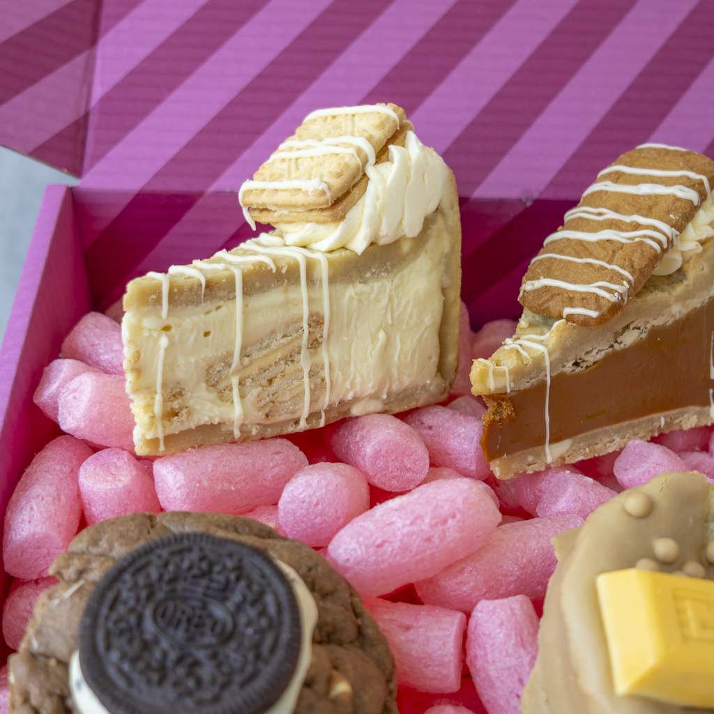 A close-up of assorted gourmet dessert slices and cookies arranged in pink bubble wrap inside a pink striped box. Visible treats include a custard cream cookie pie slice with cookie topping and drizzle, a caramel or chocolate slice with white drizzle, an Oreo cookie, and another dessert topped with a yellow square piece.
