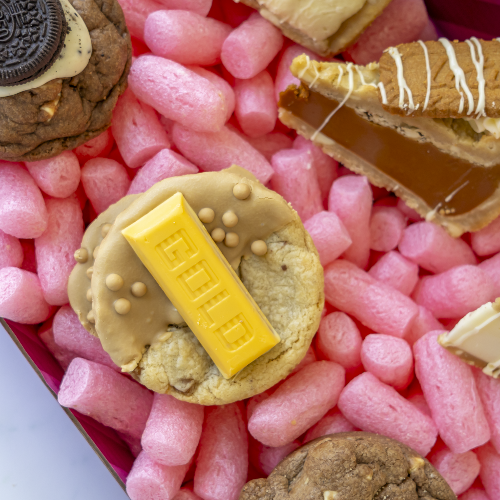 A close-up overhead view of assorted gourmet cookies arranged in a cookie box. The central cookie is topped with a yellow chocolate gold bar, surrounded by other cookies including one with an Oreo and one with Lotus biscuit and white drizzle.