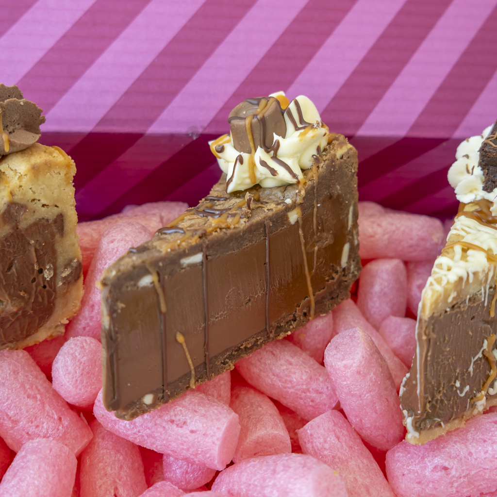 A close-up of a chocolate-covered cookie pie slice, topped with whipped cream and caramel drizzles, inside a pink striped box.