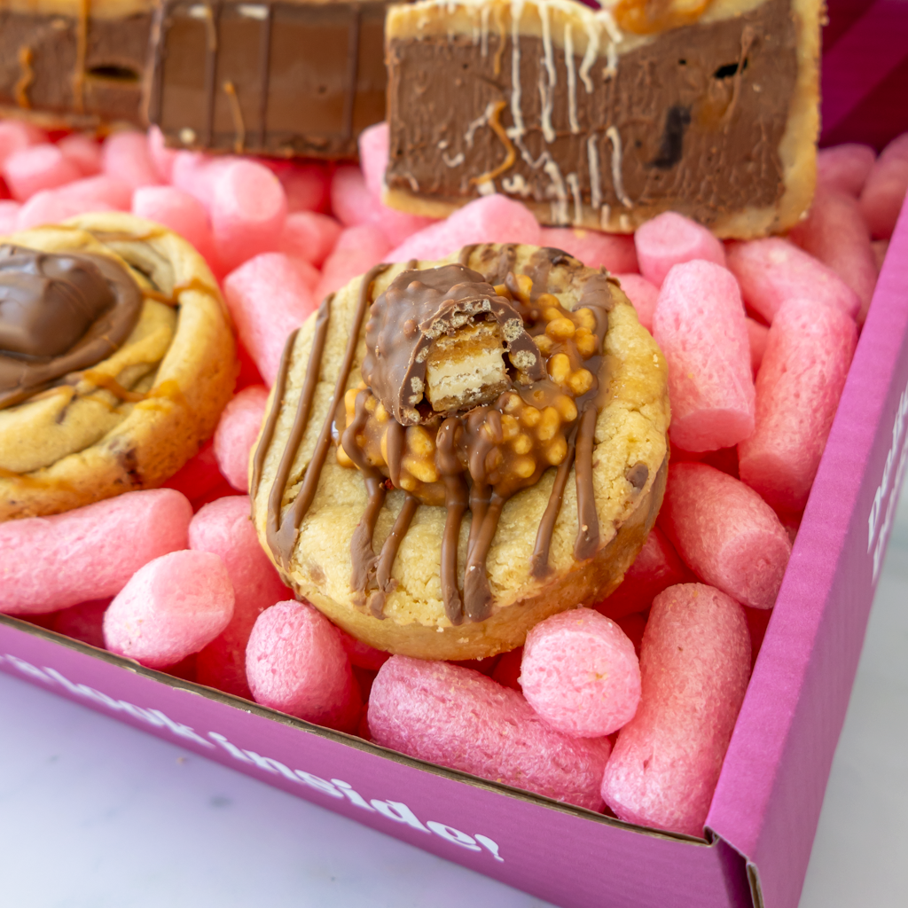 A close-up of assorted chocolate-covered cookies and a bar of chocolate arranged in pink cookie box.