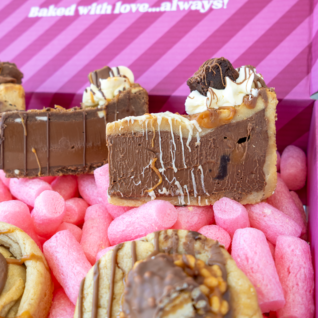 Chocolate-covered dessert bars and cookies arranged in a pink box. The box lid has the text 'Baked with love...always'