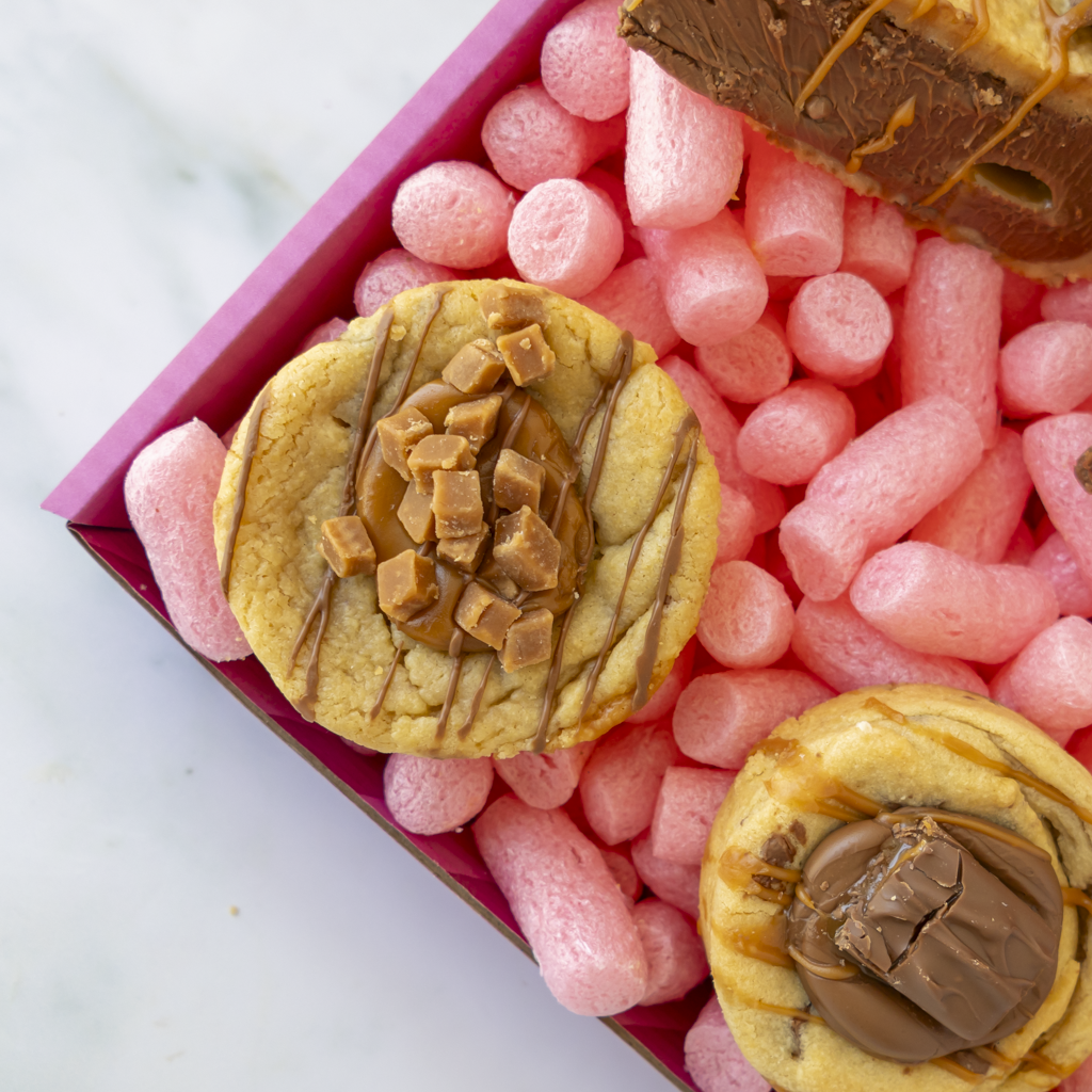 Two decorated cookie bites in a pink cookie box, with a caramel chocolate-covered cookie partially visible in the top right corner.