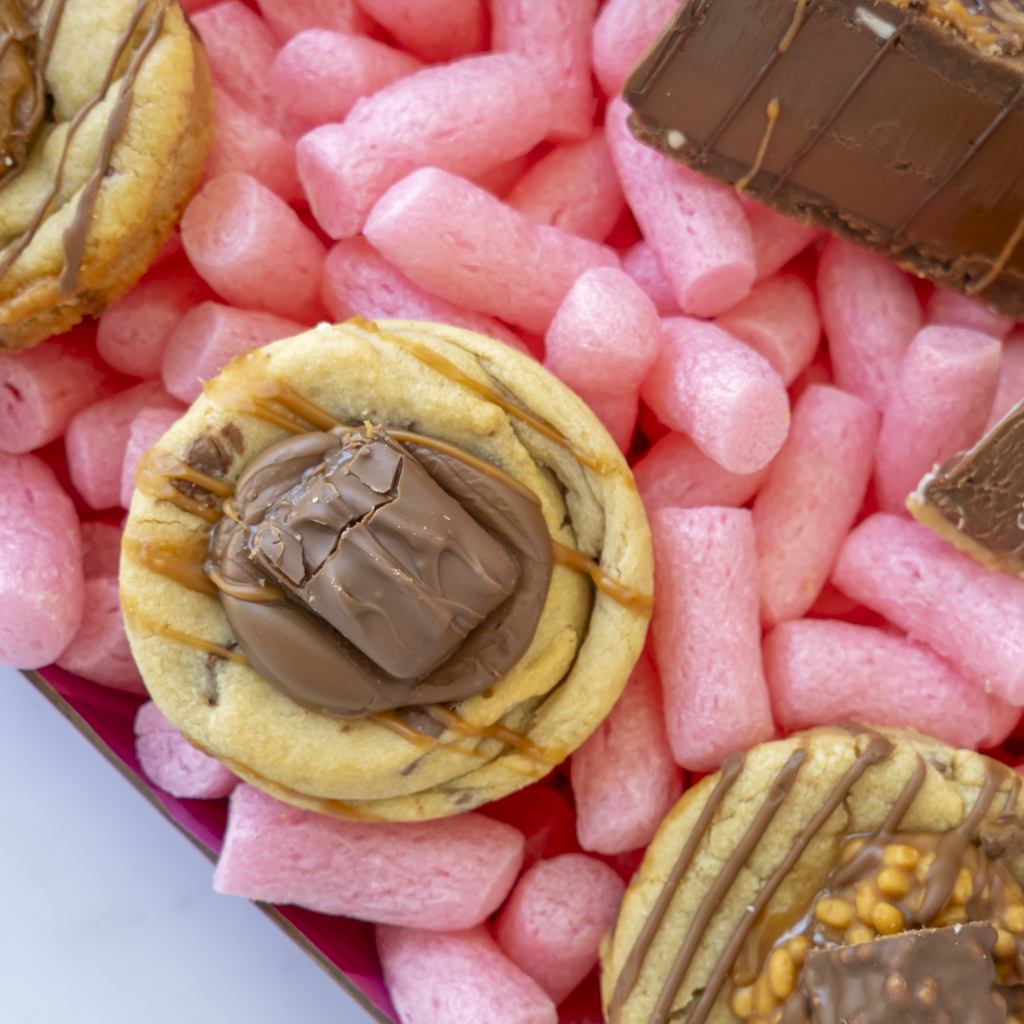 A close-up overhead view of assorted chocolate-topped cookies arranged in a box. One central cookie is topped with a large chocolate bar, and other cookies show drizzles of chocolate and caramel with nuts or caramel swirls.