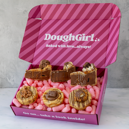 An open pink DoughGirl baking box filled with assorted chocolate- and caramel-covered pastries and cookies, surrounded by pink candy-coated pieces, on a light marble surface against a gray wall.