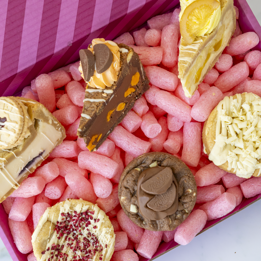 A close-up overhead view of assorted cookie pie slices and cookies arranged in a pink box. The desserts include chocolate cookie,white chocolate cookie, lemon cookie, and chocolate-covered pastry pieces.