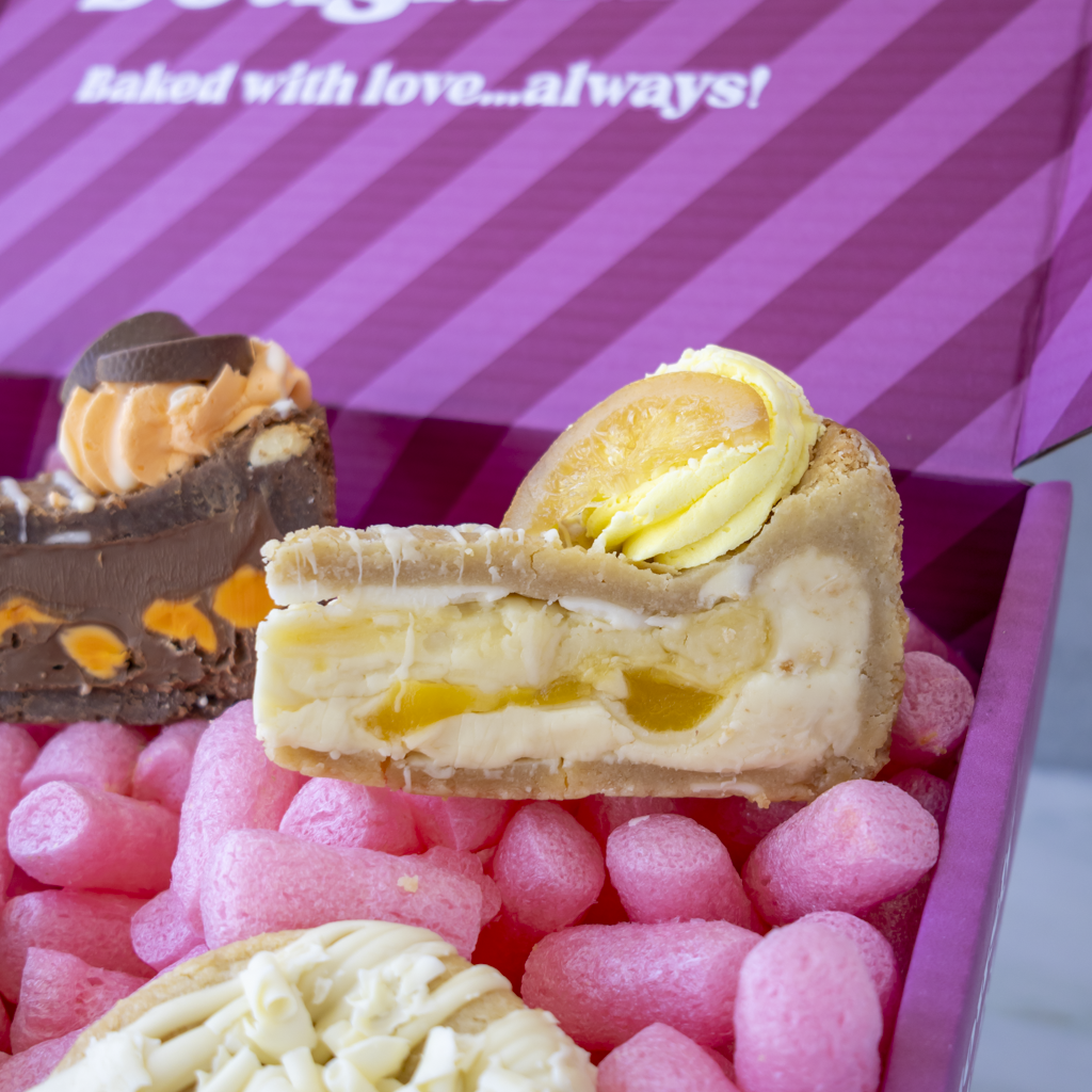 A close-up of assorted gourmet pastries arranged in a pink box. The center pastry is a cream-filled cookie pie slice with a yellow topping, and other pastries are partially visible in the background and foreground.